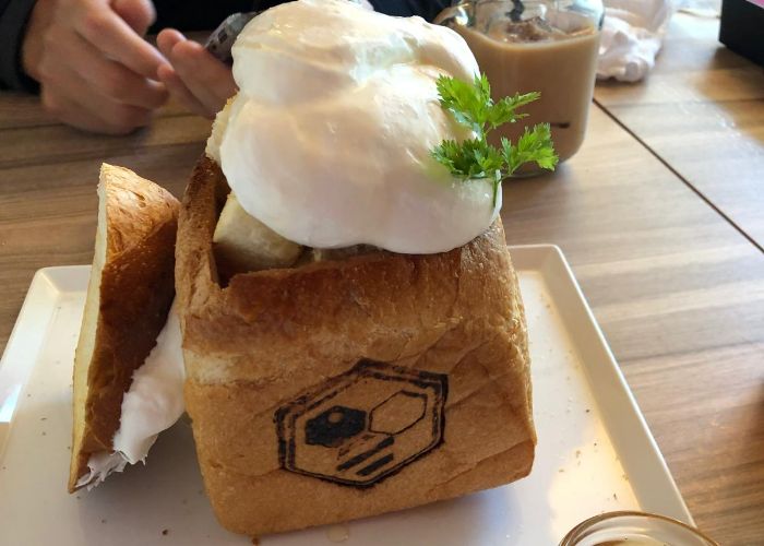 A loaf of bread has been hollowed out and filed with cubes of toasted bread and fresh cream. On the front is the Honey Toast Cafe logo of a bee.